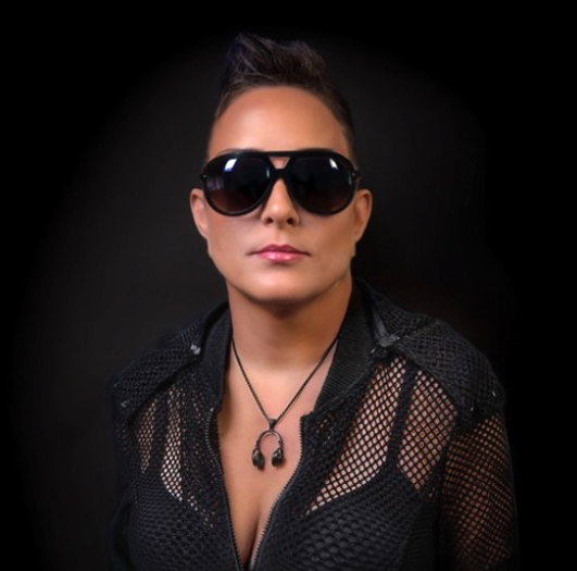 Person wearing large black sunglasses and a black mesh jacket, facing the camera against a dark background.