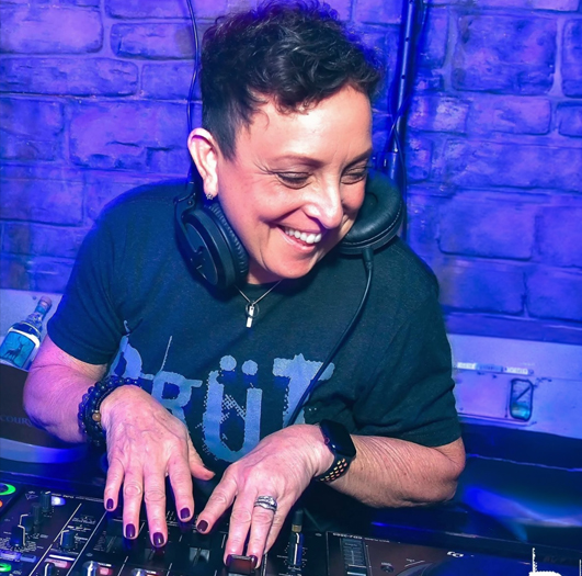 Female DJ smiling while adjusting mixer with headphones on, in a blue-lit club setting against a brick wall.