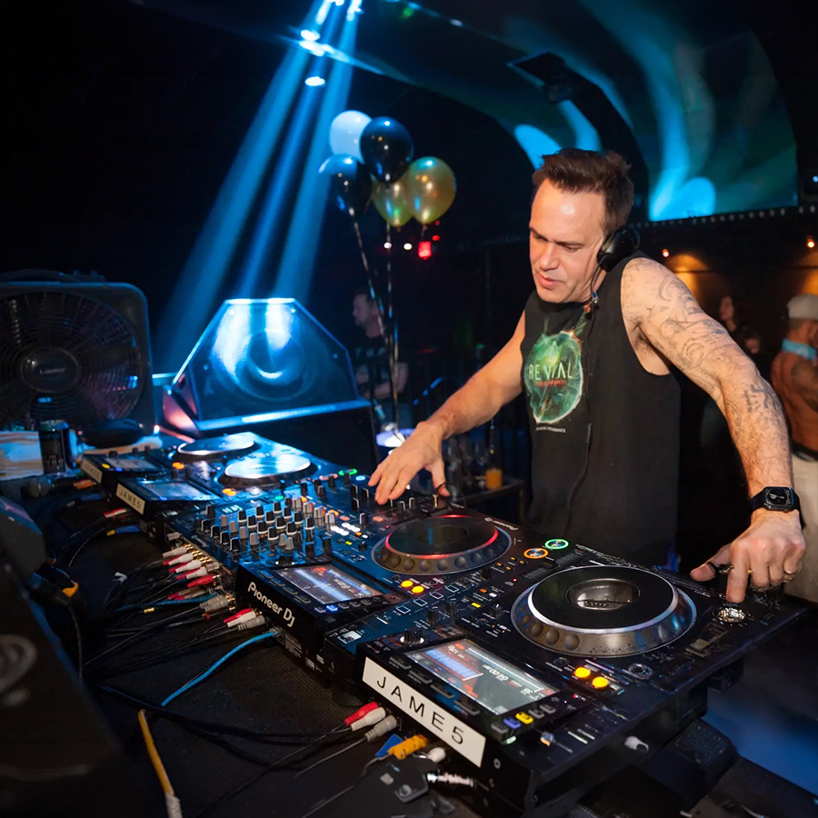 DJ wearing headphones and sleeveless shirt spins records at a club, with a mixer and turntables in front of him.