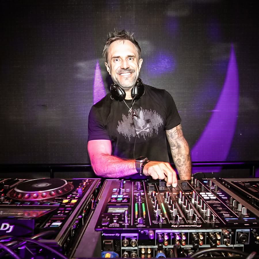 DJ wearing headphones standing behind a mixing console, performing with purple stage lighting in the background